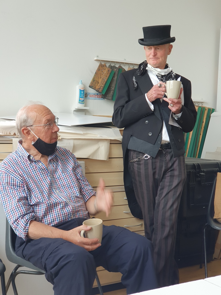 Two white men in conversation in the Highland Print Studio, holding mugs of tea. One is dressed quite casually, sitting down. The other man stands, wearing a steam-punk suit and top hat. 